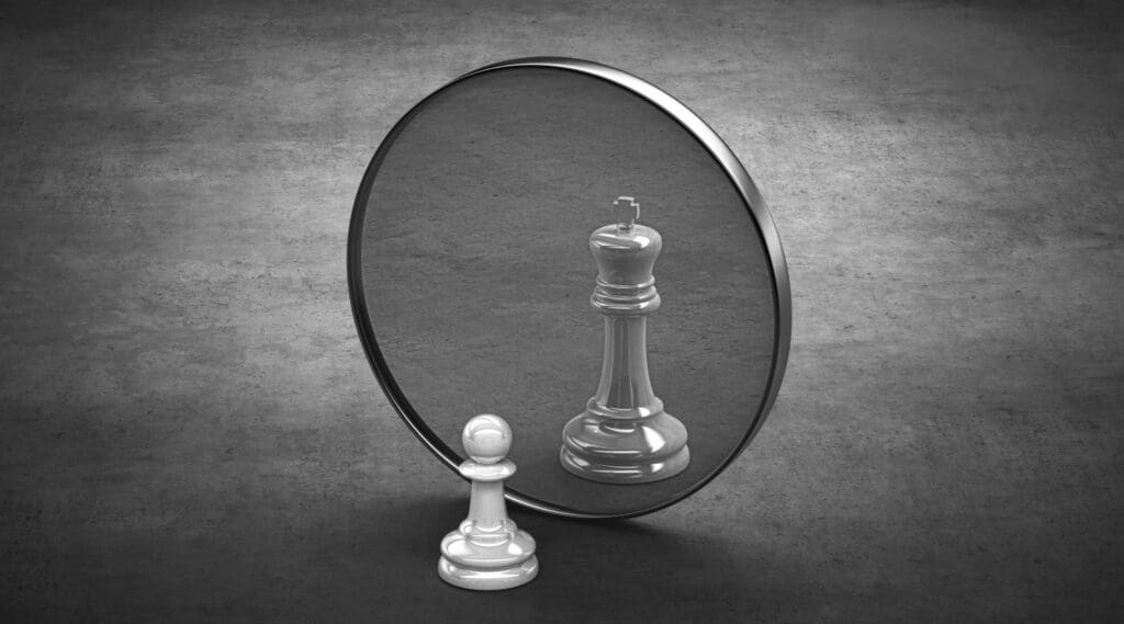 A photograph of a pawn chess piece looking at its reflection in the mirror, which appears as a king chess piece, symbolizing self-esteem. Ginny Estupinian assists individuals in enhancing their self-esteem.